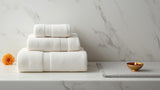 Folded off-white premium cotton towel set beside a soft silver-grey bath rug, styled for a festival-ready guest bathroom
