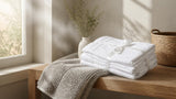 Folded white cotton towel set and grey bath rug in a sunlit bathroom, Willow and Fern premium cotton bath linen