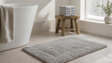 Silver grey cotton bath rug on a pale tiled bathroom floor beside a tub on a winter morning