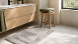 Silver grey cotton bath rug on a bright rented bathroom floor beside a wooden stool with a folded sage green towel