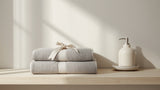 Two folded grey cotton towels tied with a ribbon on a light wooden bathroom shelf beside a soap dispenser