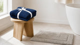 Navy blue cotton towel set on a wooden stool beside a silver grey bath rug in a bright bathroom